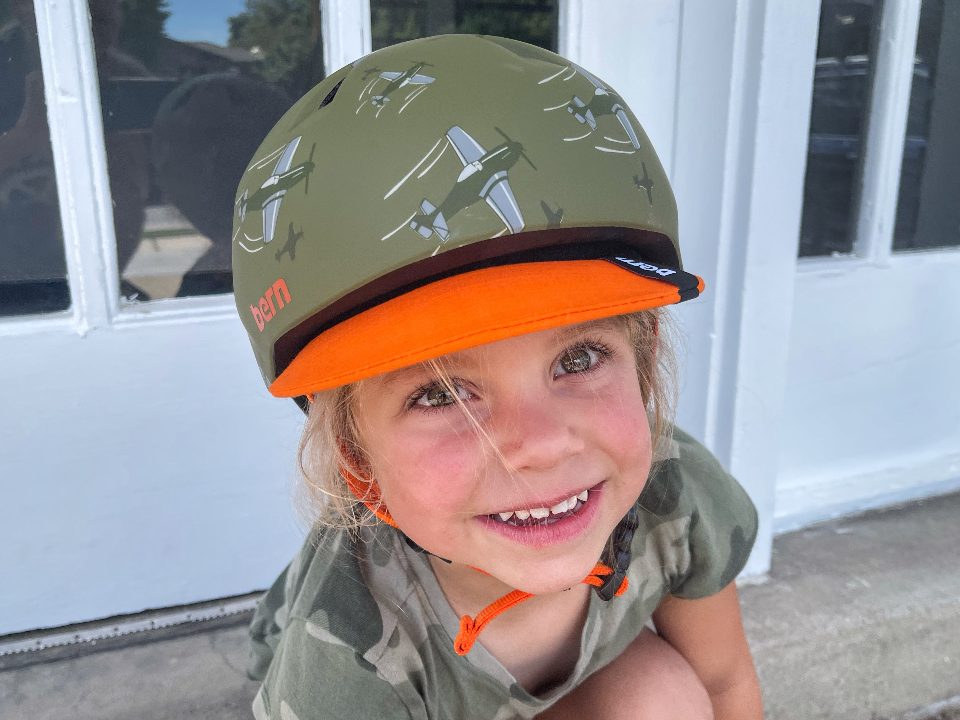 Young girl smiling and wearing the Bern Nino in army green with airplanes