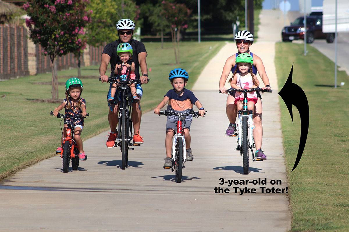 Family riding bikes - mom, dad, and two older kids on pedal bikes, with one toddler on the Tyke Toter riding with mom