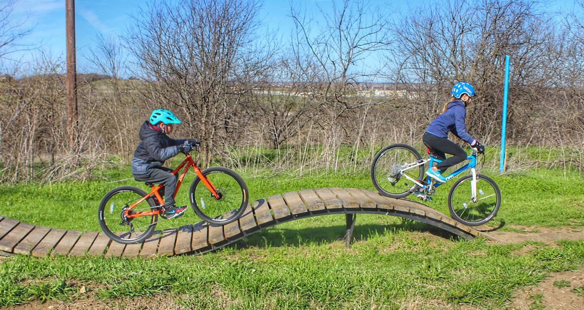 8-year-old riding Pello Reyes 24" kid's bike across wooden bridge at bike skills park, along with 10-year-old on woom 5