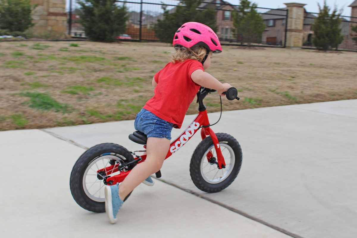 Toddler riding Ridgeback Scoot balance bike on the sidewalk. Shot from side while she's sprinting.