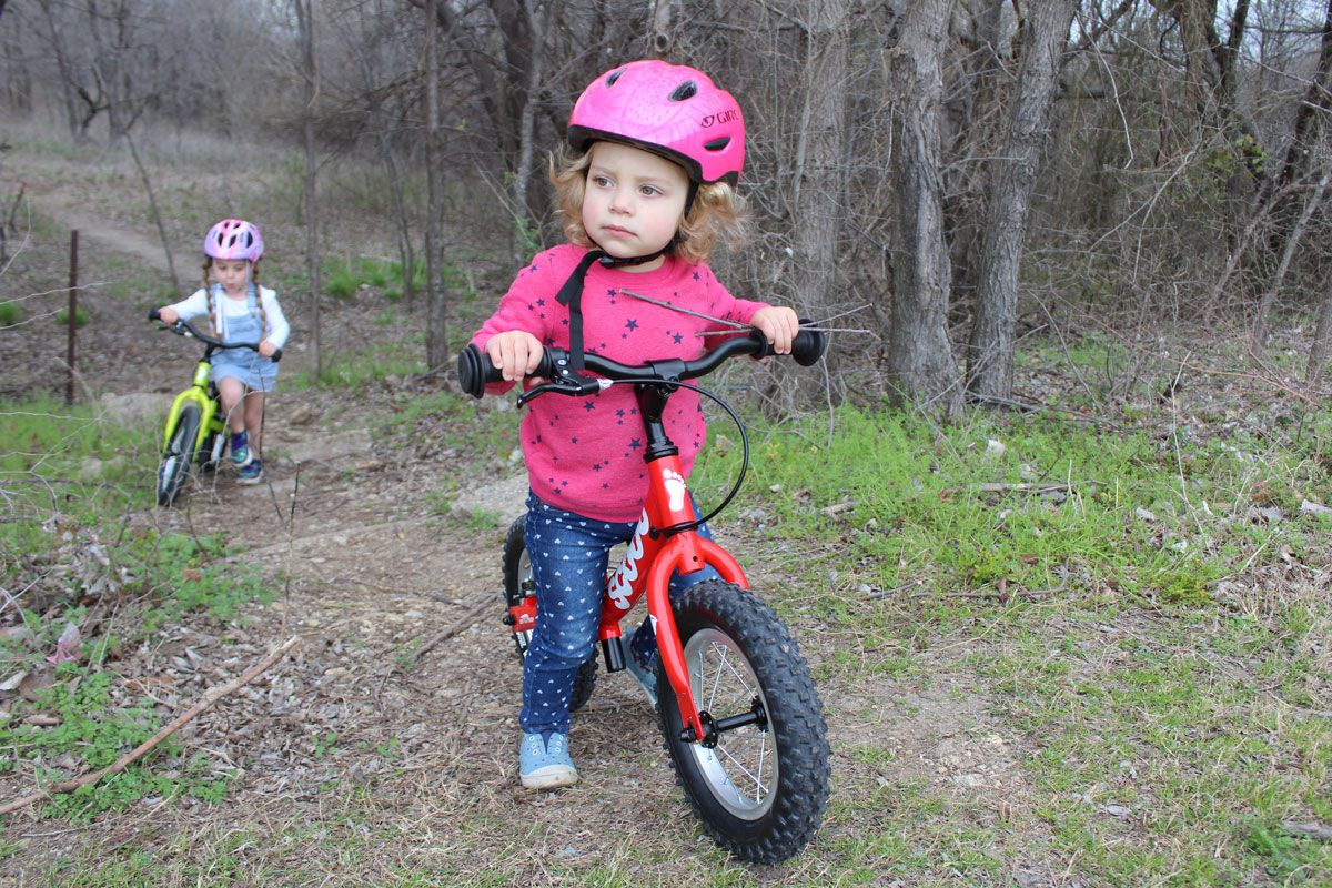 Toddler riding Ridgeback Scoot balance bike through the forest on a trail. Older toddler on Ridgeback Scoot XL behind her.
