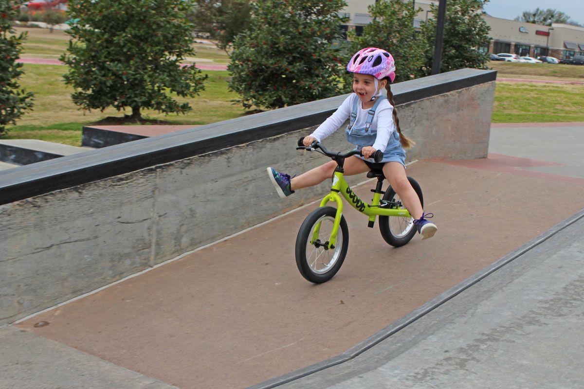 4 year old riding green Ridgeback Scoot XL balance bike at skatepark, gliding down ramp
