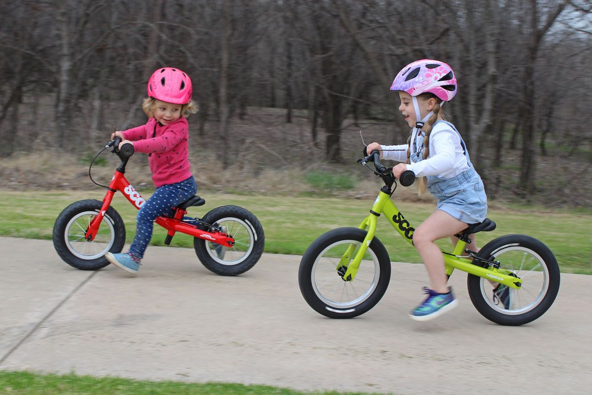Toddler riding Ridgeback Scoot and Ridgeback Scoot XL balance bikes down the sidewalk.