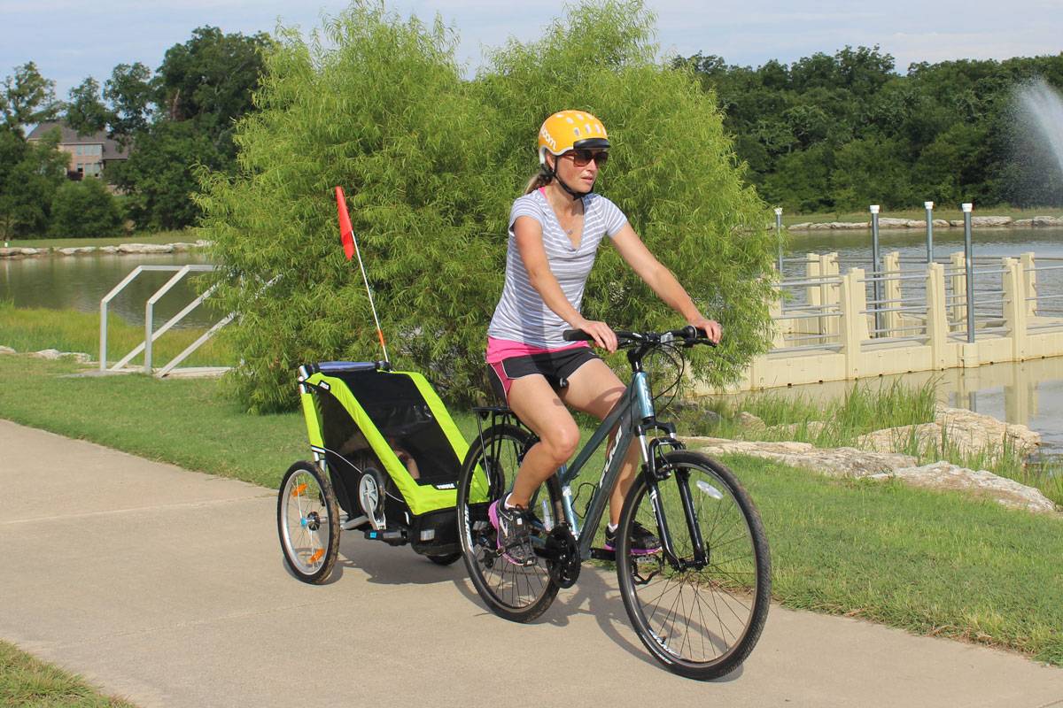 Mom pulling Thule Chariot Cheetah XT single bike trailer on paved trail by lake