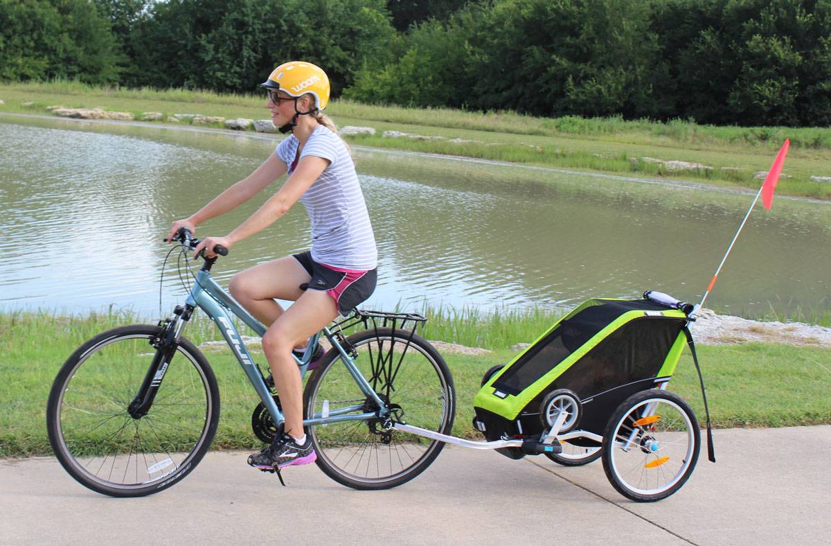 Mom pulling Thule Chariot Cheetah XT single bike trailer on paved trail by lake
