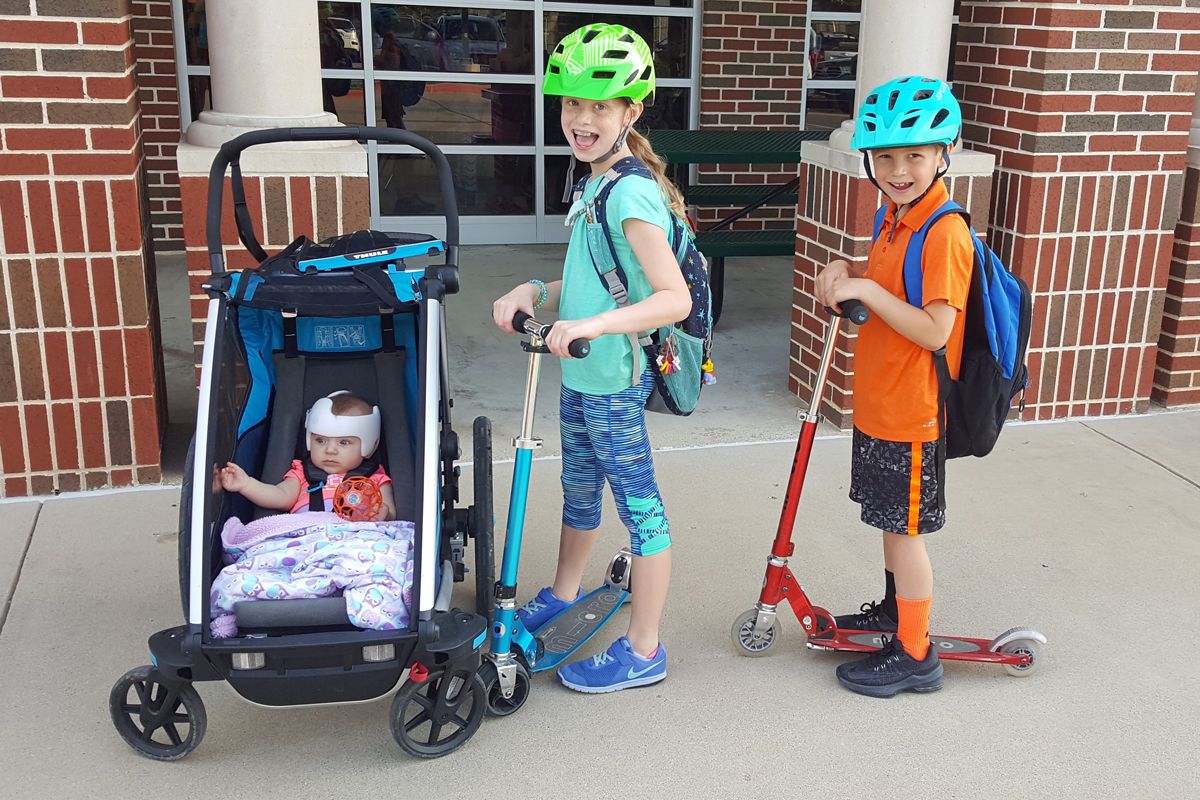 Baby sitting in Thule Infant Sling in the Thule Cross in stroller mode, next to brother and sister on Micro scooters.