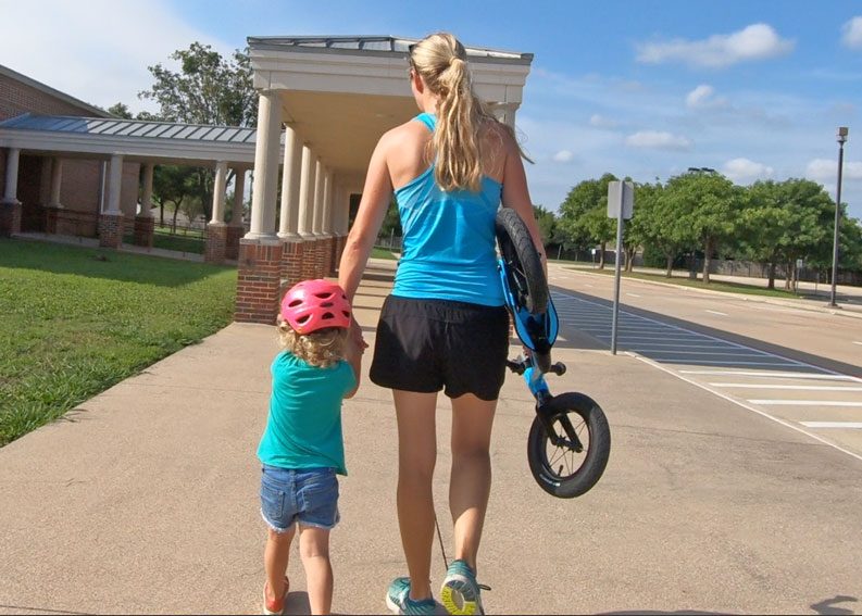 Mom carrying Vitus Smoothy balance bike while holding her toddler's hand