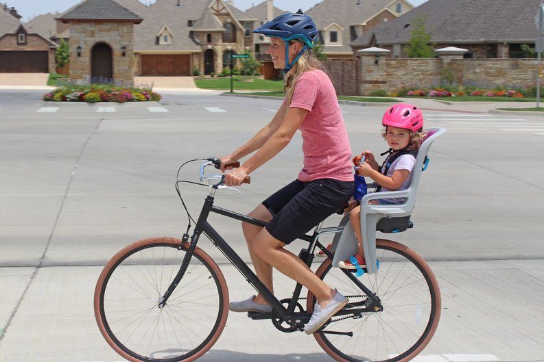 Mom riding Priority bicycle with toddler on the back in Thule RideAlong child bike seat, crossing an intersection.