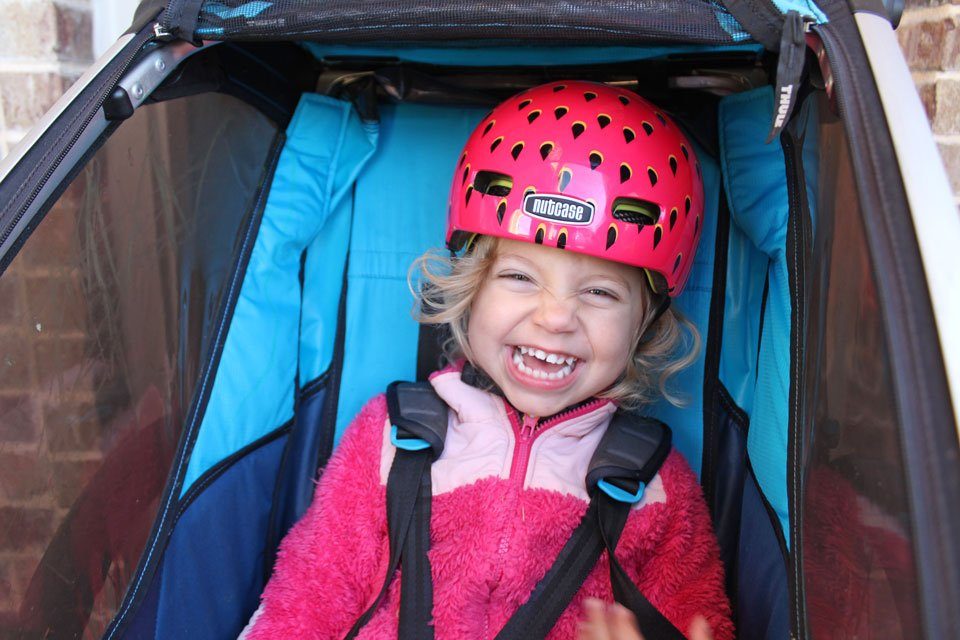 Toddler in a bike trailer, laughing and wearing the Nutcase Baby Nutty helmet that looks like a Strawberry.