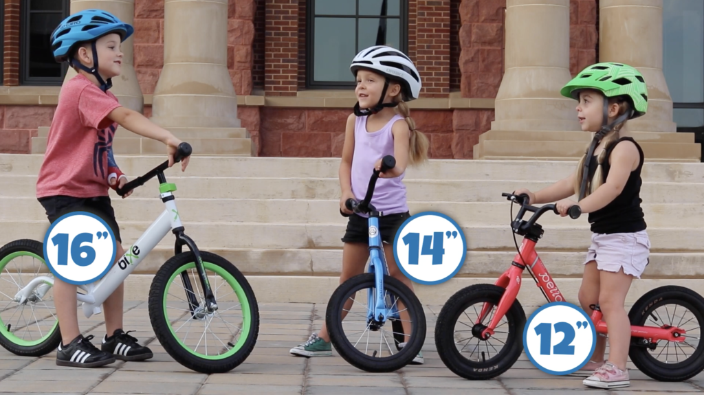Three kids, each on a balance bike. On has 16" wheels, one has 14" wheels, and one has 12" wheels