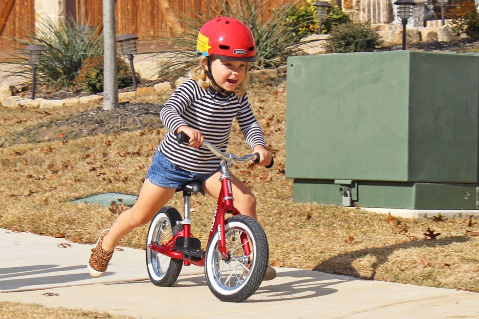 3 year old riding red Kazam bike down the sidewalk.