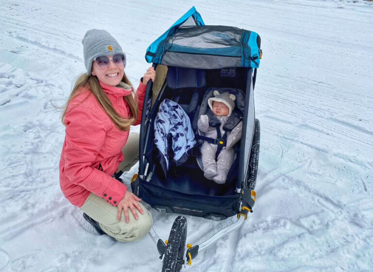 Baby in a Burley D'Lite X trailer, sitting in the Burley Baby Snuggler. Mom is standing next to him and smiling.