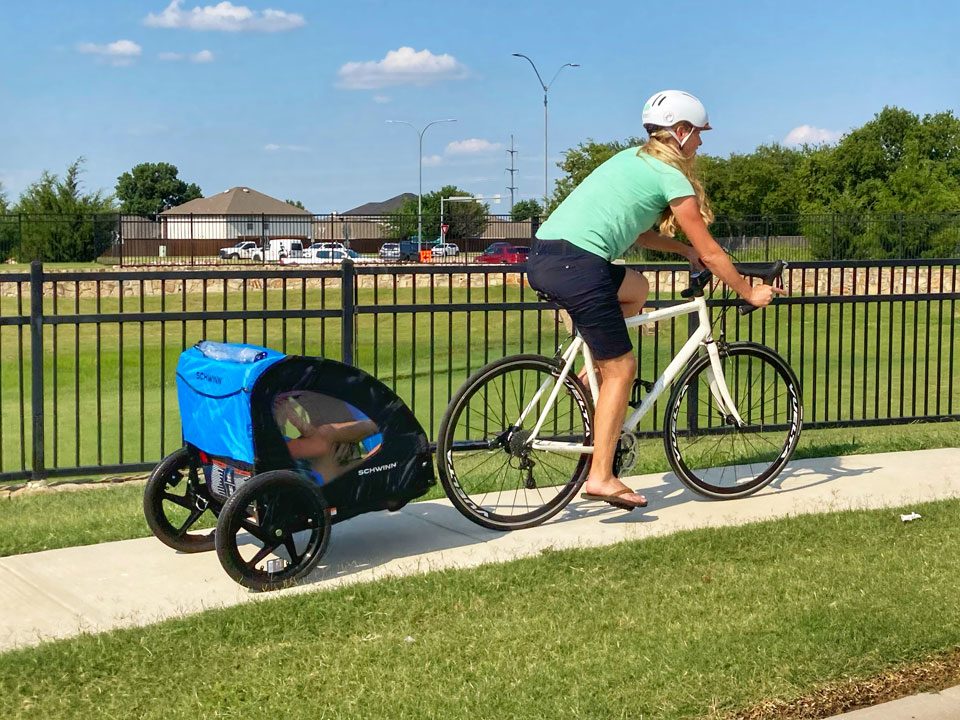 Mom riding bike with child in Schwinn Shuttle bike trailer, from behind