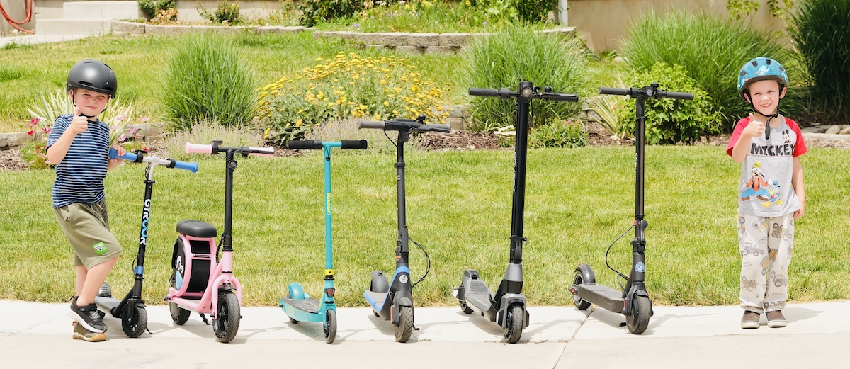 two kid testers standing next to six different electric scooters